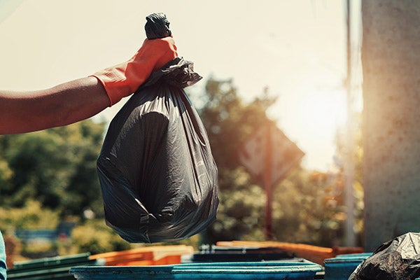 Hand holding black garbage bag above trash can.